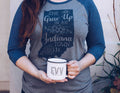 A person wearing a heather gray and navy blue raglan t-shirt with a graphic that reads 'SHE Grew Up in an Indiana Town' in chalk-style lettering. The person is holding a white enamel mug with 'E.