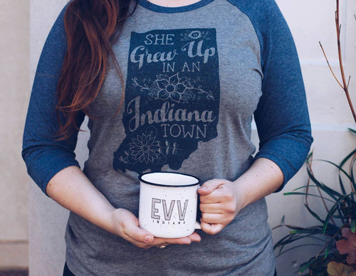 A person wearing a heather gray and navy blue raglan t-shirt with a graphic that reads 'SHE Grew Up in an Indiana Town' in chalk-style lettering. The person is holding a white enamel mug with 'E.