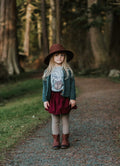 A young girl stands on a gravel path in a forest wearing a wide-brimmed brown hat, a dark green cardigan, a burgundy skirt, a graphic t-shirt, beige knit tights, and brown boots.
