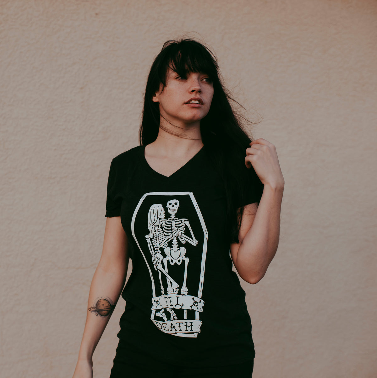 A woman with dark hair wearing a black V-neck t-shirt with a white graphic of a skeleton in a coffin and the words 'TIL DEATH'. She is standing against a textured beige wall.