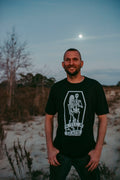 A man wearing a black t-shirt with a white coffin graphic and the text 'TIL DEATH' stands outdoors in a sandy, dry landscape at dusk under a full moon.