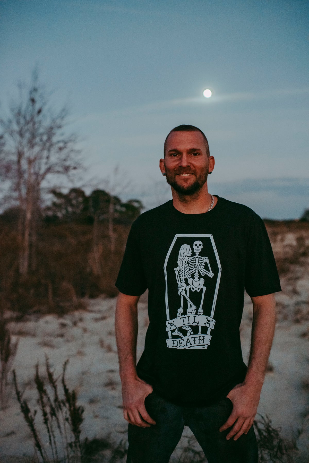 A man wearing a black t-shirt with a white coffin graphic and the text 'TIL DEATH' stands outdoors in a sandy, dry landscape at dusk under a full moon.
