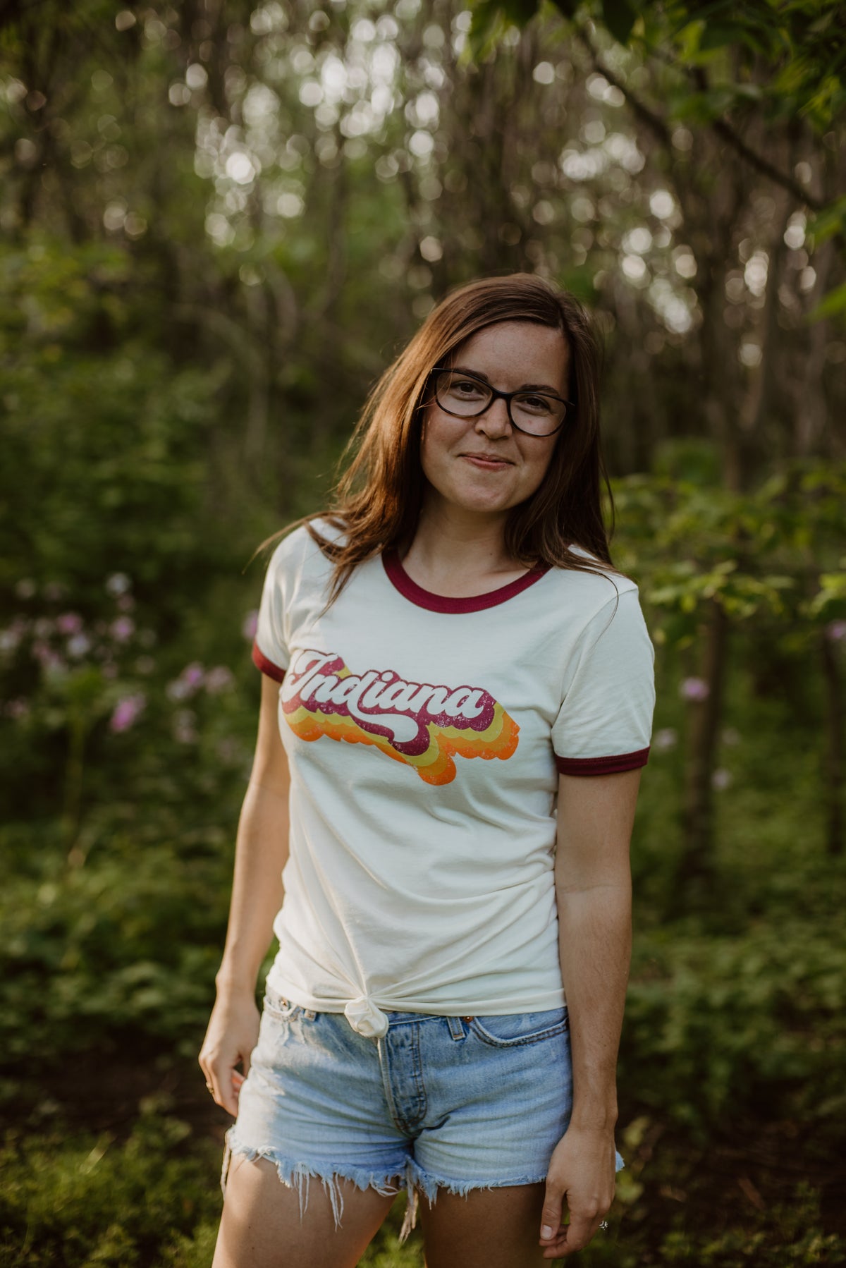 A woman wearing a white ringer T-shirt with a maroon crew neck and sleeve trim, featuring a retro-style 'Indiana' graphic in orange and yellow. She is also wearing denim shorts and glasses, standing in a sunlit forest setting.