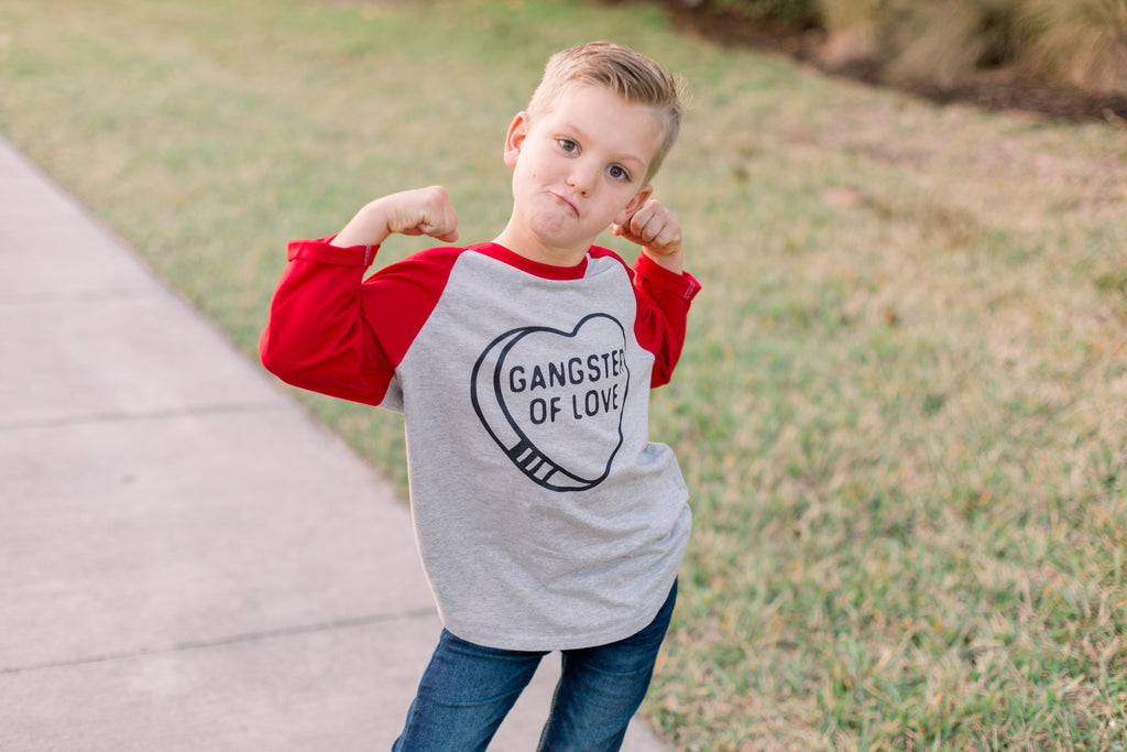 A young boy posing outdoors on a concrete path next to a grassy lawn, wearing a heather gray and red raglan t-shirt with a black heart graphic that reads 'GANGSTER OF LOVE'.