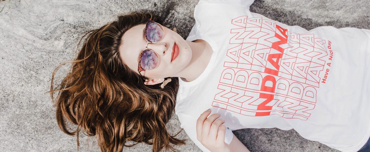 A woman lying on a gray concrete surface wearing a white graphic t-shirt with 'INDIANA INDIANZ' printed in red and pink, along with pink-tinted sunglasses.