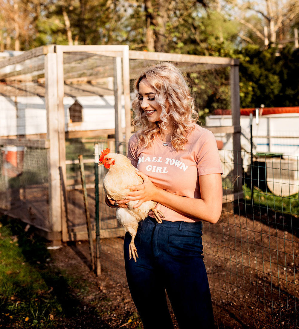 A woman standing in a backyard with a chicken coop holding a white chicken. She is wearing a pink t-shirt with the text 'SMALL TOWN GIRL' and dark blue jeans.