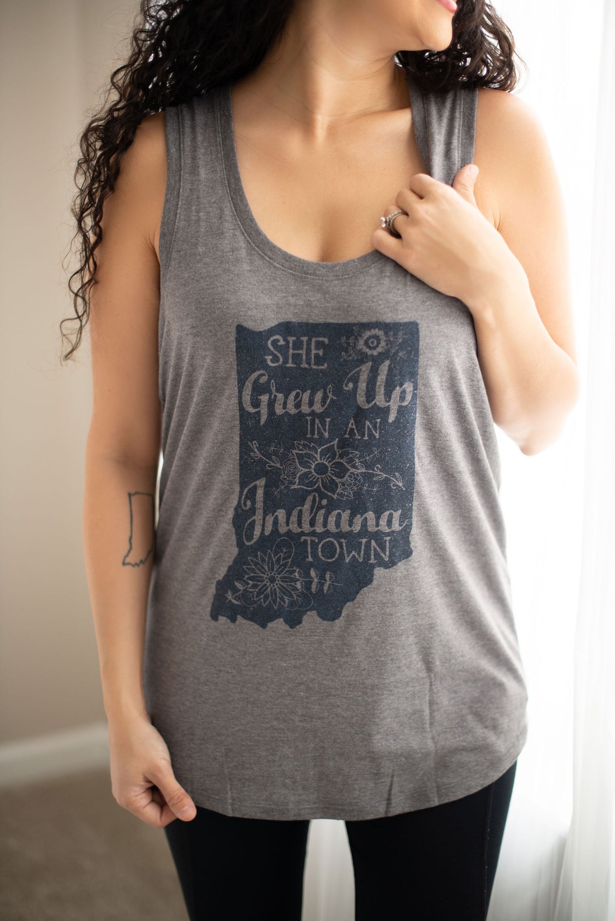 A woman wearing a heather gray sleeveless tank top with a dark navy graphic of the state of Indiana and the text 'She Grew Up in an Indiana Town'.