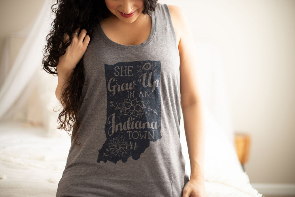 A woman wearing a heather gray sleeveless tank top with a dark navy graphic of the state silhouette of Indiana and the text 'SHE Grows Up IN An Indiana TOWN' in a bedroom setting.