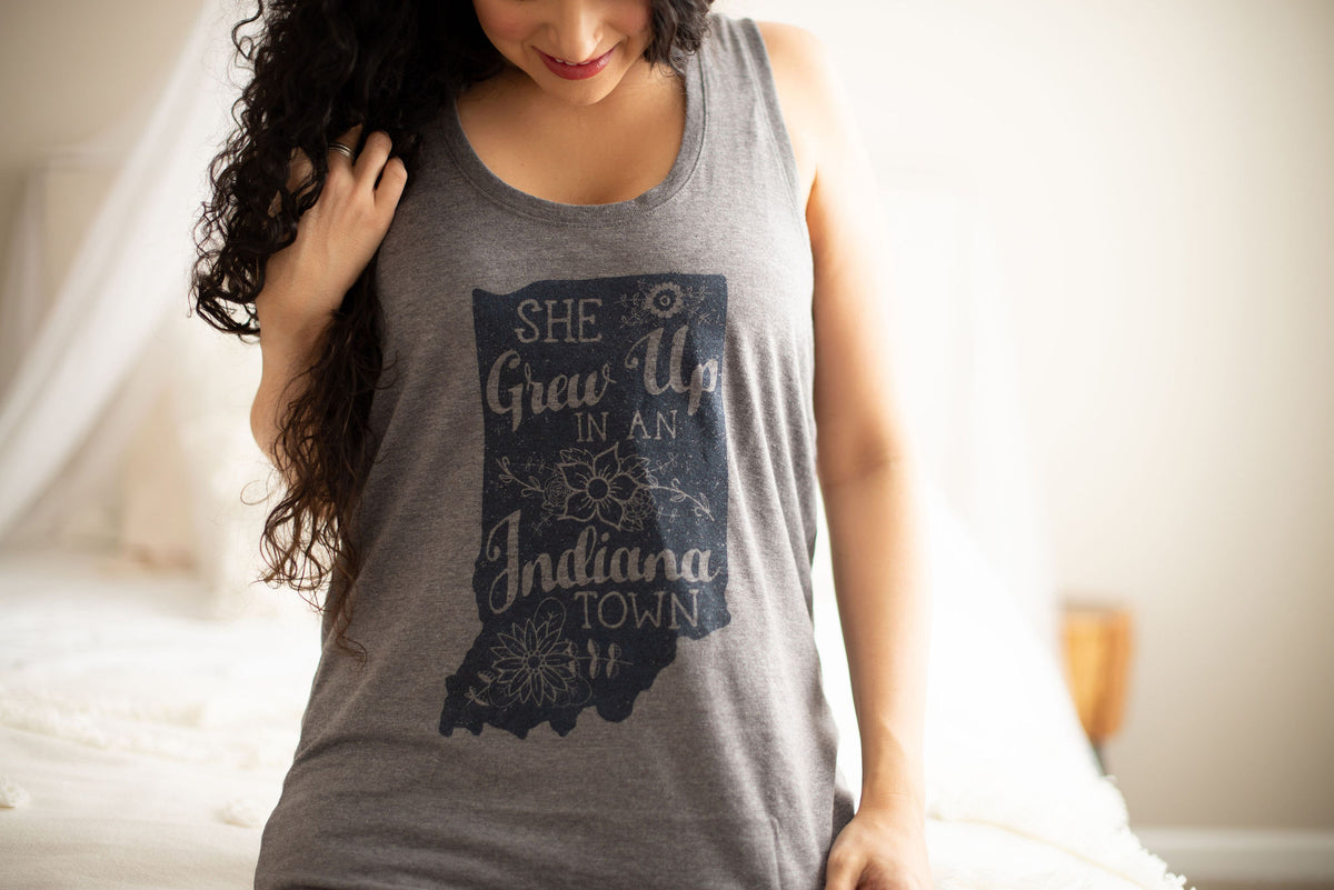 A woman wearing a heather gray sleeveless tank top with a dark navy graphic of the state silhouette of Indiana and the text 'SHE Grows Up IN An Indiana TOWN' in a bedroom setting.