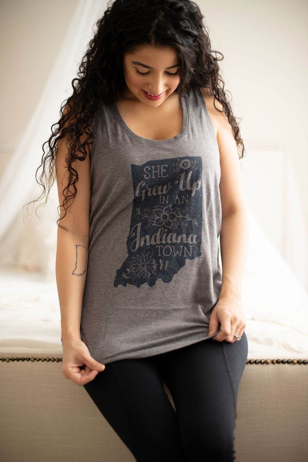 A woman wearing a heather gray sleeveless tank top with a dark navy graphic of the state of Indiana and the text 'She Grew Up in Indiana Town', paired with black leggings, sitting on a beige upholstered bench.