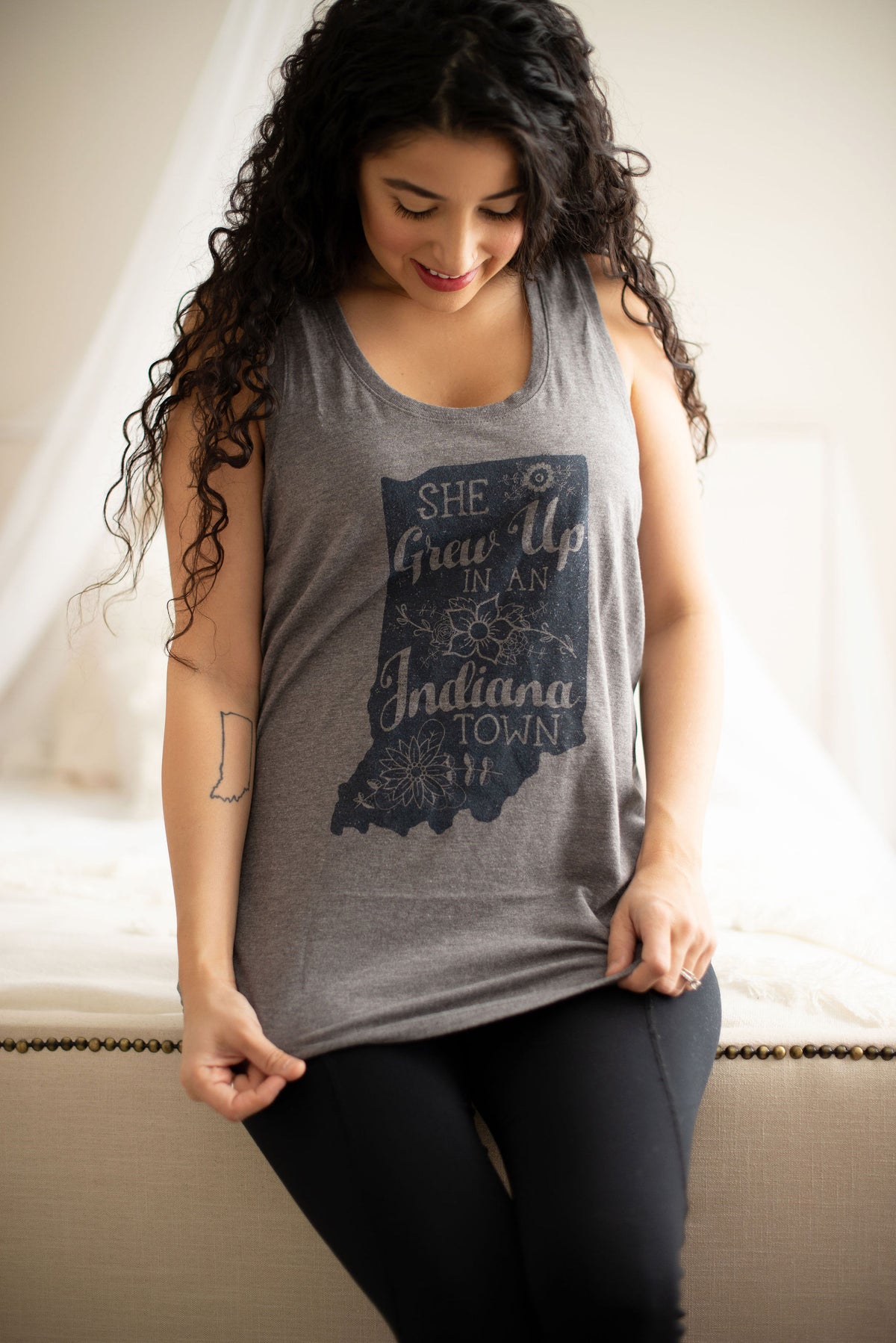 A woman wearing a heather gray sleeveless tank top with a dark navy graphic of the state of Indiana and the text 'She Grew Up in Indiana Town', paired with black leggings, sitting on a beige upholstered bench.
