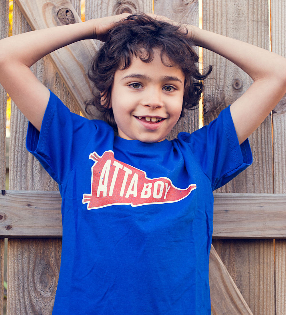 A child wearing a royal blue t-shirt with 'Atta Boy!' text and a red and white pennant design.